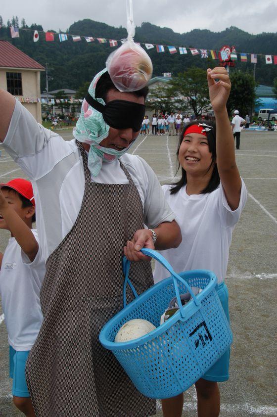 宮宿小学校かぼちゃ運動会の競技「親子でかぼちゃりんご狩り」に参加中のかぼちゃを持った青いかごを持つ目隠しをしたエプロン姿の男性と男児児童と目の前に吊るされたりんごに手を伸ばす女児児童の写真