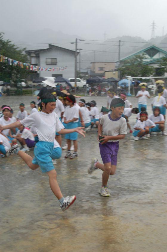リレーの最中に降り出した大雨の中水浸しになった校庭で緑色のハチマキをしたかぼちゃのバトンを手渡した女児児童とバトンを受け取り駆け出す同じく緑色のハチマキをした男児児童の写真