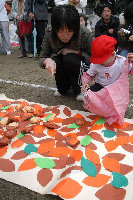 さゆり保育園の園庭でピンク色のエコバッグに紙でできた食べ物を拾い入れる園児と指を差して指示をする母親の写真