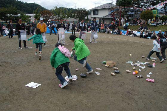 さゆり保育園の園庭で競技「森をきれいにしよう」に参加しゴミを拾いゴミ袋を持っている男性たちに向かって走っている地元の小学生たちの写真