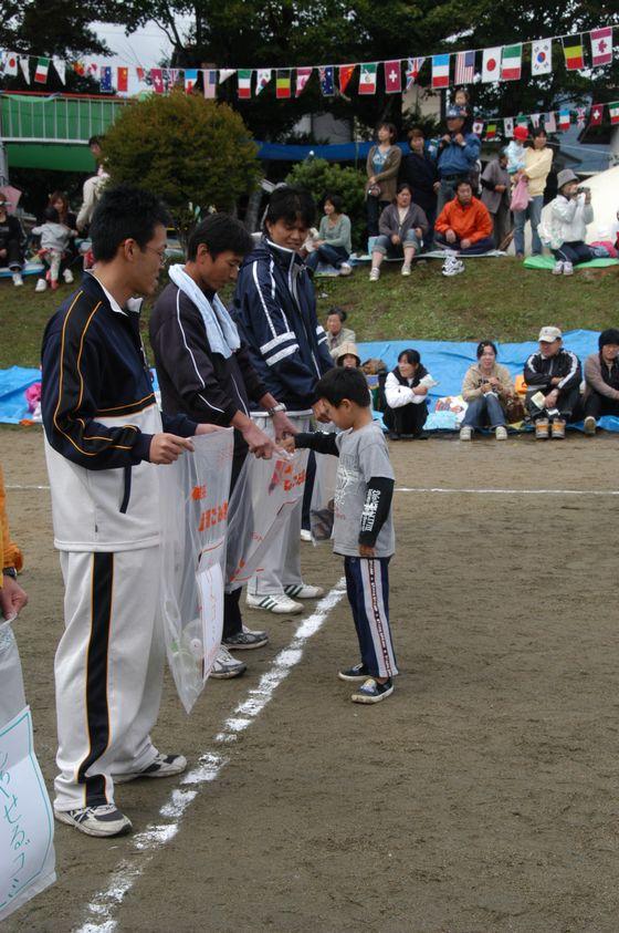 競技「森をきれいにしよう」で拾った園庭のゴミを男性たちが持つゴミ袋に分別して捨てる地元の男児小学生の写真