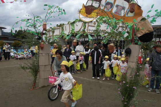 さゆり保育園の園庭で「親子競技 地球にやさしいお買い物」に参加するとなりのトトロの猫バスが添えられたエコリンピックの文字が書かれた入場門をくぐる黄色いエコバッグを持った女の子の園児とピンク色の子ども用自転車を引く母親と入場門でスタートを控えている親子たちの写真