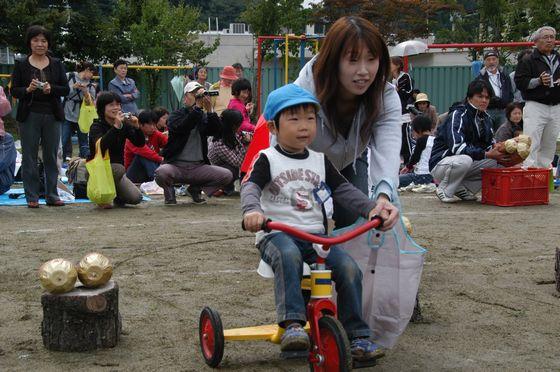 「親子競技 地球にやさしいお買い物」で三輪車に乗ってゴールインした男の子の園児と白いエコバッグを左手首に提げながら三輪車のハンドルを握る男の子の左手を握る母親の写真