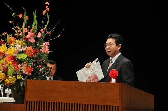 近藤洋介衆議院議員が壇上で紙面を広げ、賞賛の言葉を送っている写真