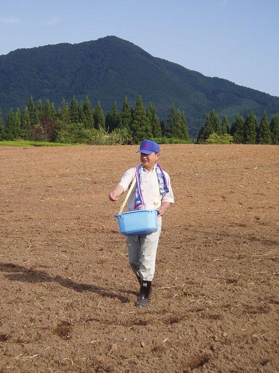 畑の上で肩に提げた青いかごに入った菜の花の種を蒔く副町長の写真