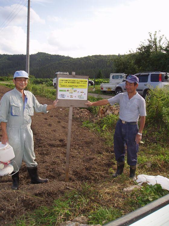 菜の花の種が蒔かれた畑に立てた看板を片手で支え合い笑顔を浮かべる2人の男性の写真