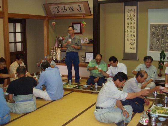 収穫祭の会場で乾杯の挨拶をする男性とビールの乾杯をする「菜の花の種まき・収穫祭」の参加者たちの写真