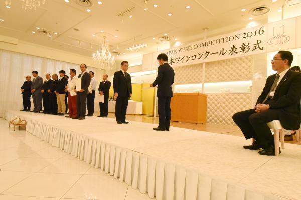 山梨県甲府市にあるホテルベルクラシック甲府で開かれた「日本ワインコンクール」の表彰式の壇上で賞状の受け渡しを行う2人の男性と賞状を受け取った他の受賞者と壇上で着席している出席者の写真
