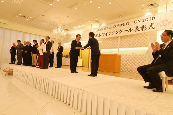 日本ワインコンクール2016表彰式の壇上で賞状を受け取る鈴木浩幸町長と登壇したワイナリーの代表者たちと拍手をする壇上で着席している男性の写真