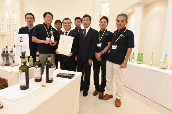 日本ワインコンクール2016の会場で賞状を持ち笑顔を浮かべる鈴木浩幸町長と町長の周りにいる朝日町ワインスタッフたちの写真