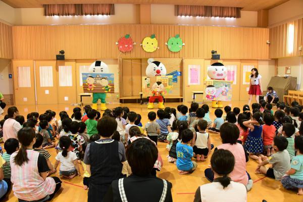 あさひ保育園で大勢の子どもや大人の前で稲作戦隊おこめんジャーが食育イベントショーをしている様子の写真