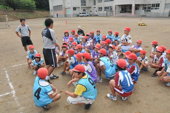 大谷小学校の校庭で行われたモンテディオ山形によるサッカー教室でスケジュールを説明する都コーチと説明を聞く教室に参加中の児童たちの写真