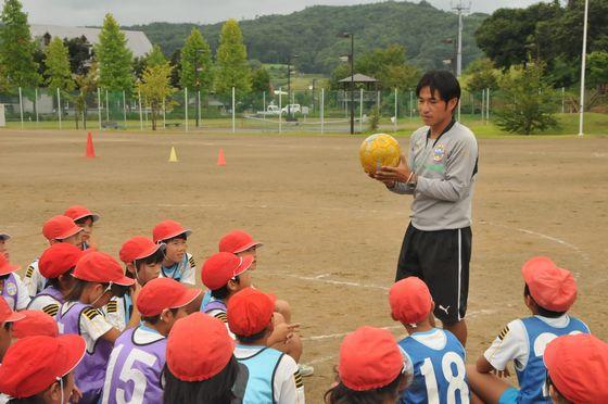 大谷小学校の校庭で黄色いボールを手にした都コーチとサッカー教室に参加中の児童たちの写真
