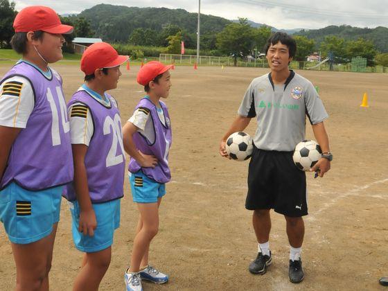 大谷小学校の校庭で2つのサッカーボールを持つ茨木コーチとサッカー教室に参加中の3人の児童の写真