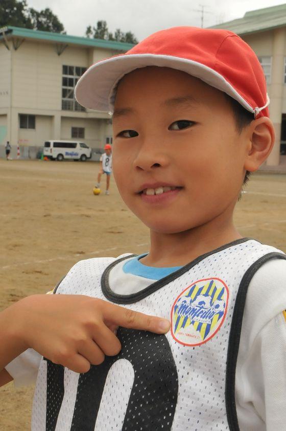 大谷小学校の校庭で着用しているゼッケンの左肩付近についているモンテディオ山形のロゴマークを指差す児童の写真