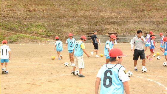 大谷小学校の校庭で生徒に指導をする2人のコーチと指導を受けるサッカー教室に参加中の児童たちの写真