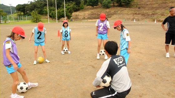 大谷小学校の校庭でコーチの指導の下両足でボールを使って練習をしている児童たちの写真