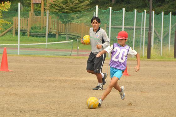 ミニゲーム中に黄色いボールを使ってドリブルをする児童と後ろで見つめる黄色いボールを抱えている都コーチの写真