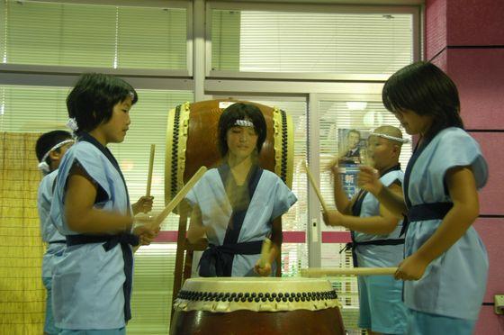 豊年太鼓を披露する宮宿小学校の3人の児童と、その後ろで太鼓を叩く2人の自動の写真