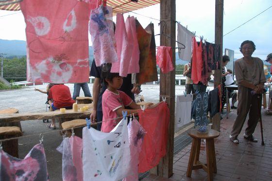 旧上郷小学校の入り口付近で作品の染物を乾かすために吊るし干しをしている女子児童とその様子を見つめる杖をついている中高年女性の写真