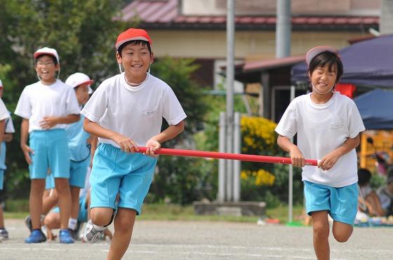 2人で赤い棒を持ち合いながら校庭を並んで走る男児の生徒の写真