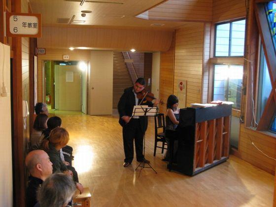 旧立木小学校の廊下で開催された「ジプシーヴァイオリンのコンサート」でバイオリンを弾く男性とピアノを弾く女性と演奏を聴く人々の写真