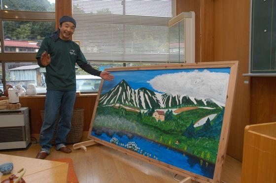 旧立木小学校の教室で両手を広げそばにある自身が手掛けた絵画を紹介する長岡昂司さんの写真
