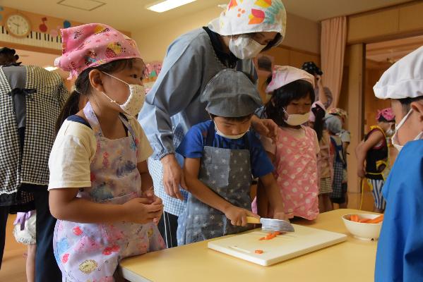 園児が先生に見守られながらニンジンを切っている写真